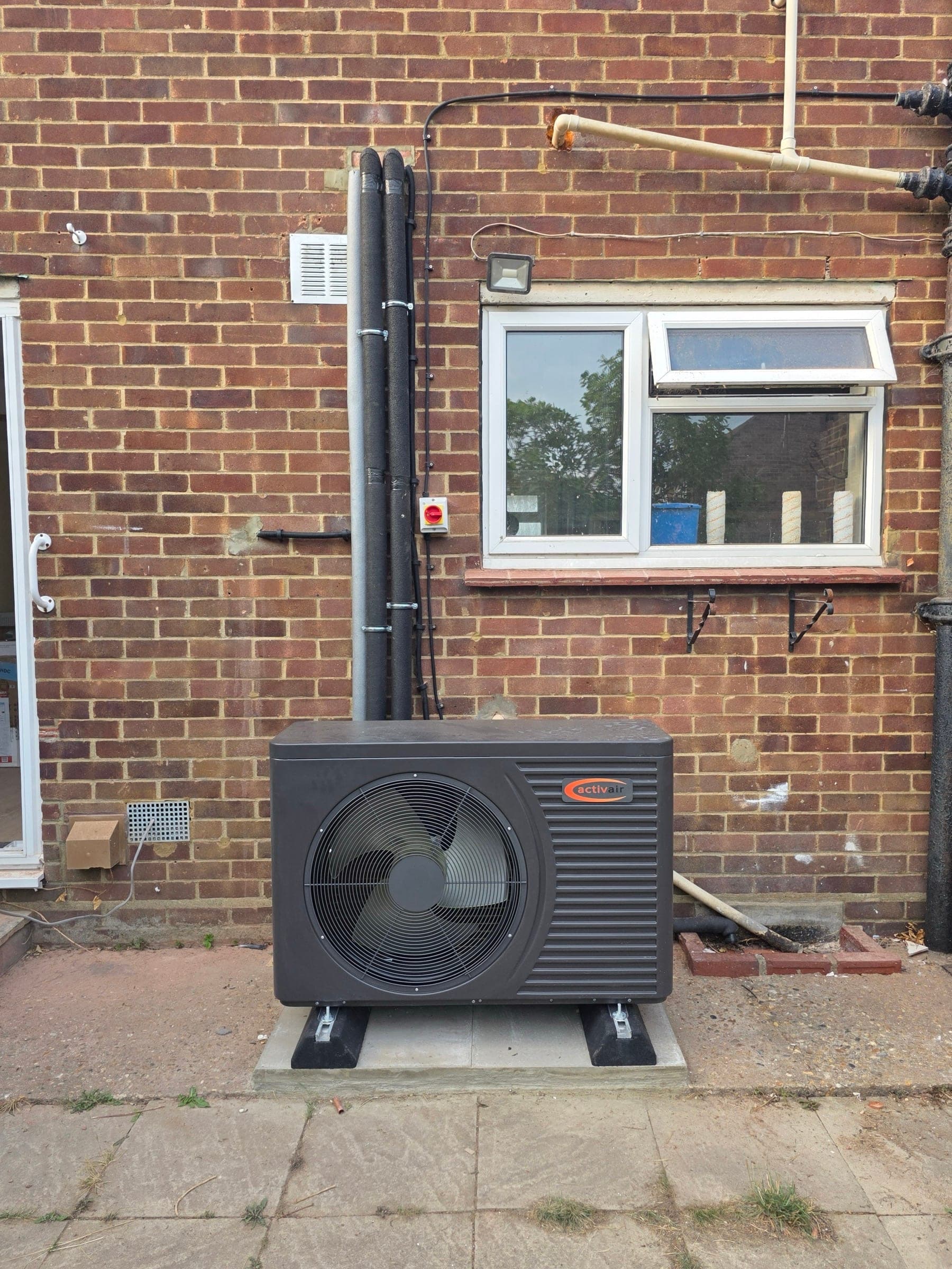 An activair air source heat pump installed against the rear wall of a brick house, sitting level on anti-vibration feet on a concrete pad, with insulated flow and return pipework running up the wall and an isolator switch fitted alongside.