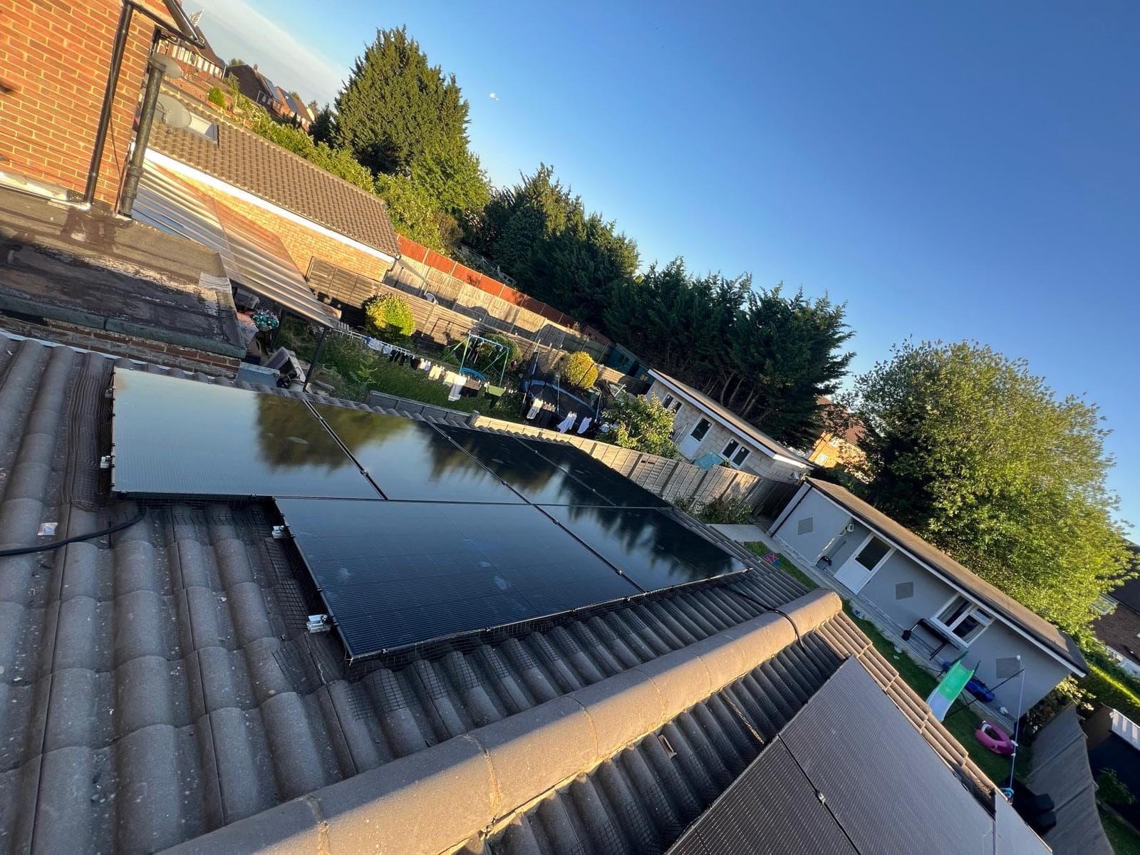A completed all-black solar photovoltaic array on a tiled pitched roof, with bird protection mesh clipped around the edge of the panels and a garden and neighbouring houses beyond.